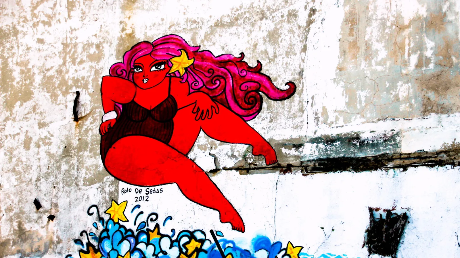 Gemaltes Wandbild einer roten Frau mit pinken Locken und einem Seestern im Haar, die auf blauen Wellen mit gelben Sternen sitzt, auf einer verwitterten weißen Wand.
