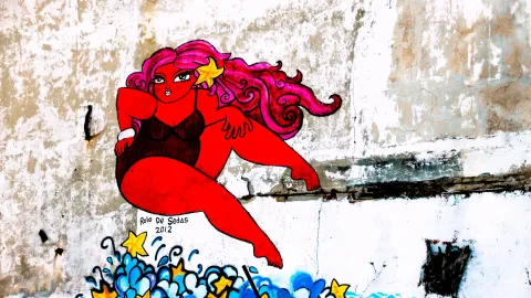 Gemaltes Wandbild einer roten Frau mit pinken Locken und einem Seestern im Haar, die auf blauen Wellen mit gelben Sternen sitzt, auf einer verwitterten weißen Wand.