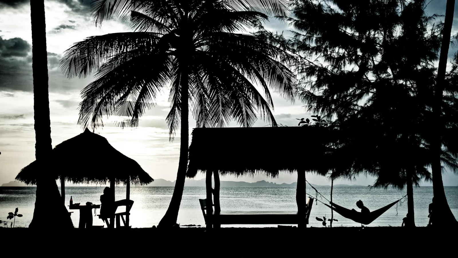 Schwarzweiß-Silhouette eines Strands mit Palmen, Strohhütten und einer Person in einer Hängematte vor einem Meer mit Inseln am Horizont