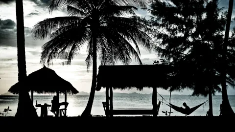 Schwarzweiß-Silhouette eines Strands mit Palmen, Strohhütten und einer Person in einer Hängematte vor einem Meer mit Inseln am Horizont