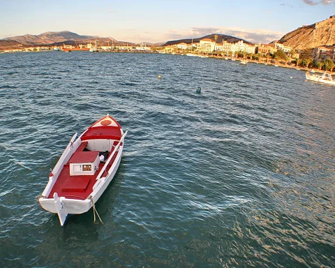 Ein rot-weißes Ruderboot liegt im Hafenbecken, im Hintergrund eine Küstenstadt mit weißen Häusern, Booten und Bergen.
