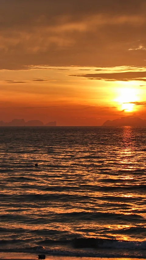 Orangefarbener Sonnenuntergang über einer welligen Meeresoberfläche, am Horizont zeichnen sich dunkle Inselsilhouetten ab.