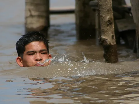 Ein Junge mit nassen dunklen Haaren schwimmt im braunen Wasser des Mekong, nur Kopf und Schultern ragen heraus, Wasser spritzt vor seinem Gesicht. Im Hintergrund stehen Holzpfähle und eine Konstruktion am Ufer.