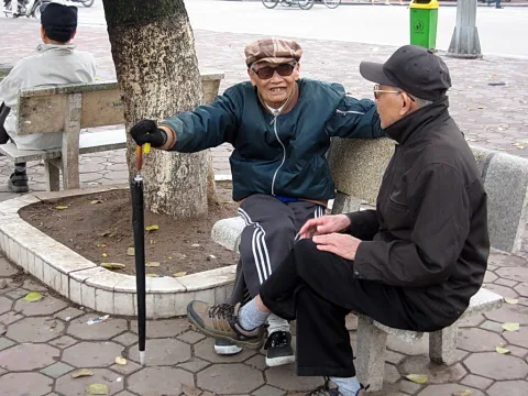 Zwei ältere Männer sitzen auf einer Steinbank neben einem Baum. Der linke hält einen Schirm oder Stock, trägt Mütze, Sonnenbrille und eine grüne Jacke. Der rechte trägt eine dunkle Kappe und eine dunkle Jacke und blickt zu ihm. Im Hintergrund stehen eine Mülltonne und ein Radfahrer auf einer Straße.