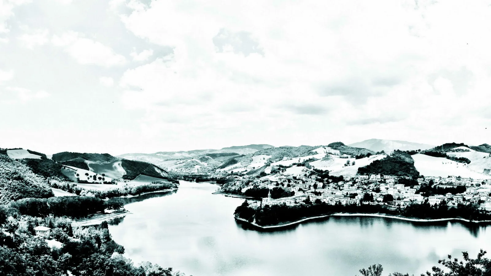 Schwarzweiß-Panorama: Ein breiter See zieht sich durch eine hügelige Landschaft, am rechten Ufer liegt ein dichtes Dorf unter einem bewölkten Himmel.