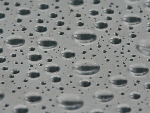 Viele Wassertropfen unterschiedlicher Größe verteilen sich auf einer grauen, glatten Oberfläche und spiegeln das Licht.