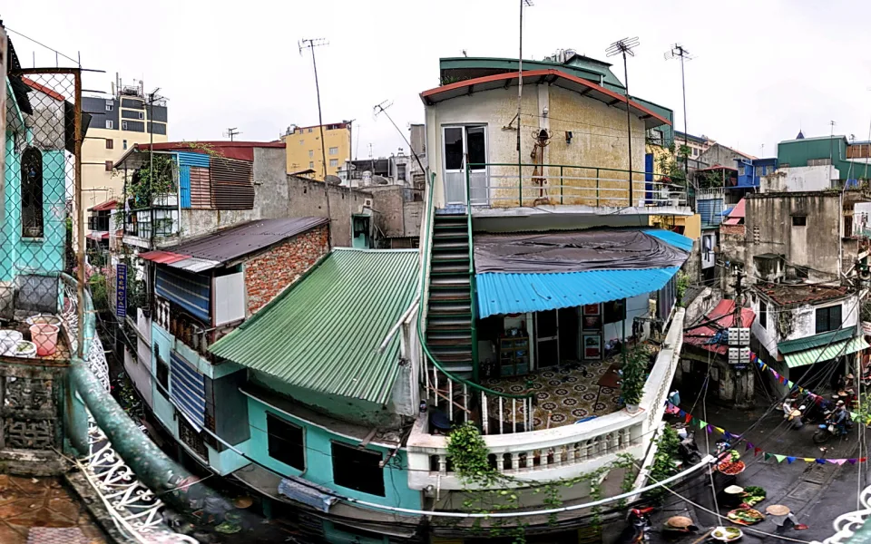 Mehrstöckige Häuser in einer engen Gasse in Hanoi. Auf einem Dach steht eine Außentreppe mit grünem Geländer, darunter spannen sich grüne und blaue Wellblechvordächer. Im Hintergrund hängen bunte Wimpelketten über der Straße, unten sitzen Menschen bei Essensständen.