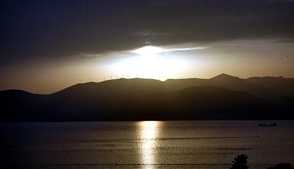 Sonnenuntergang hinter einer Bergkette am Meer, im Gegenlicht sind kleine Windkraftanlagen auf dem Kamm zu erkennen, rechts fährt ein Schiff über das Wasser.