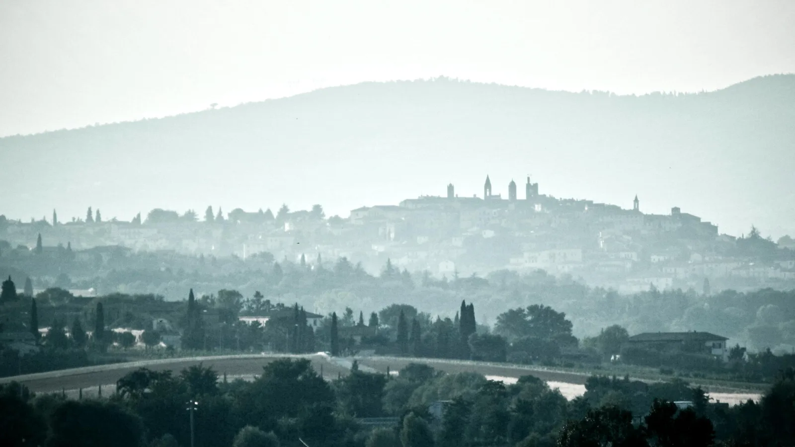 Eine Stadt auf einem Hügel, im Dunst als graue Silhouette mit Kirchtürmen, davor Baumreihen und Zypressen, dahinter ein bewaldeter Bergrücken.