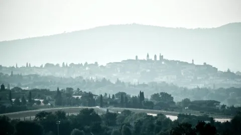Eine Stadt auf einem Hügel, im Dunst als graue Silhouette mit Kirchtürmen, davor Baumreihen und Zypressen, dahinter ein bewaldeter Bergrücken.