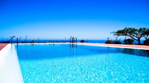 Ein türkisblaues Schwimmbecken mit silbernen Einstiegsleitern auf einer gefliesten Terrasse, dahinter Sträucher, ein Baum und das blaue Meer unter wolkenlosem Himmel.