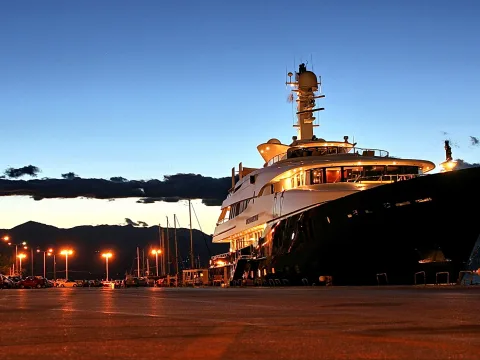 Große weiße Luxusyacht mit beleuchteten Decks liegt abends im Hafen, im Hintergrund dunkle Berge und ein Parkplatz mit Laternen.