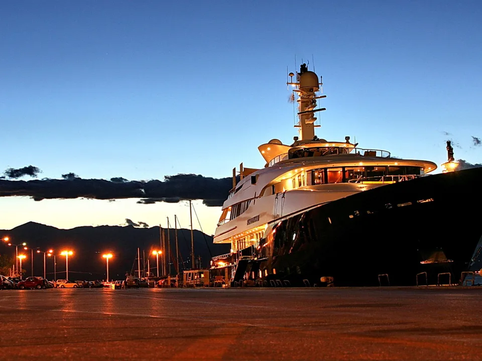Große weiße Luxusyacht mit beleuchteten Decks liegt abends im Hafen, im Hintergrund dunkle Berge und ein Parkplatz mit Laternen.