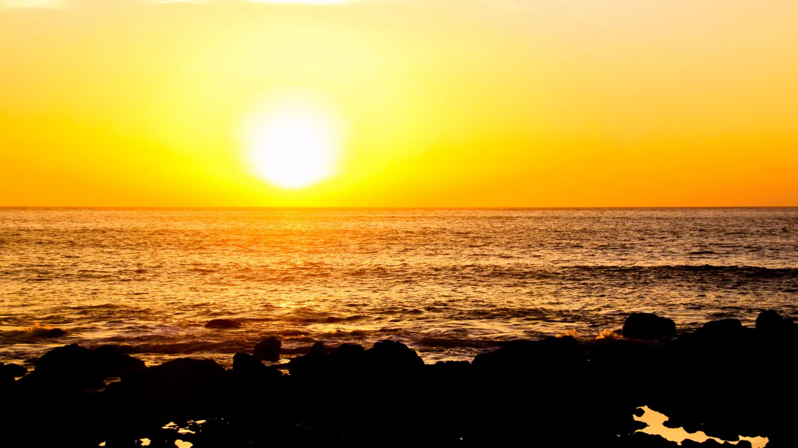 Glutorange Sonne kurz über dem Horizont, darunter eine ruhige Meeresfläche mit Wellen und dunkle Felsen im Vordergrund.