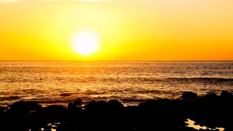 Glutorange Sonne kurz über dem Horizont, darunter eine ruhige Meeresfläche mit Wellen und dunkle Felsen im Vordergrund.