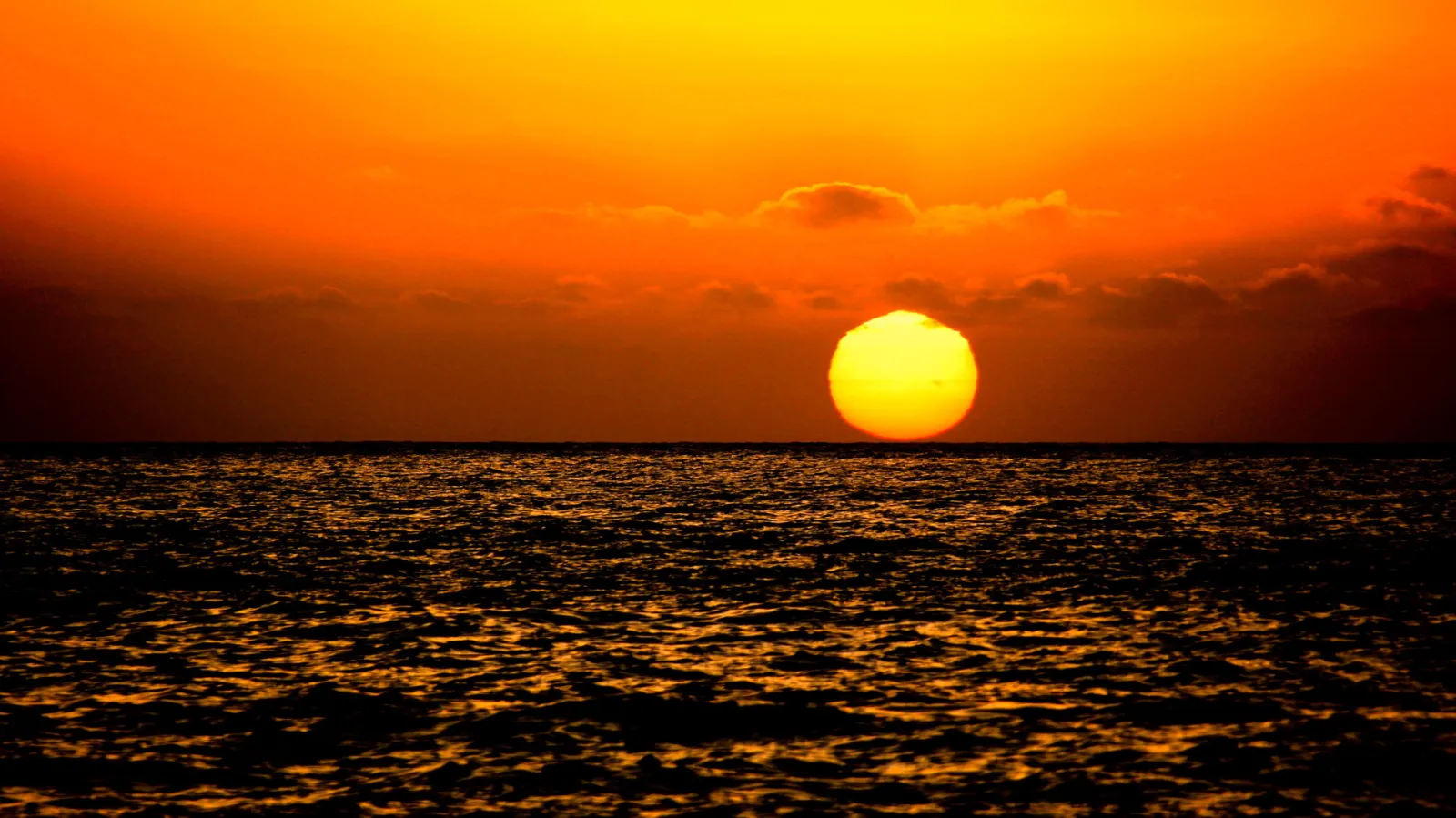 Große gelbe Sonne knapp über dem Meereshorizont, darüber orangefarbener Himmel mit dünnen Wolken, unten glitzernde Wellen.