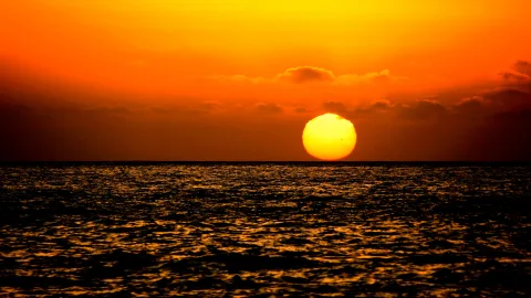 Große gelbe Sonne knapp über dem Meereshorizont, darüber orangefarbener Himmel mit dünnen Wolken, unten glitzernde Wellen.