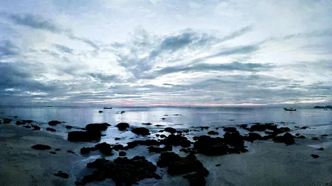 Felsiger Strand bei Dämmerung mit dunklen Steinen im Vordergrund, ruhigem Meer und einem schmalen rosa Streifen am Horizont unter bewölktem Himmel. Zwei kleine Boote liegen auf dem Wasser.
