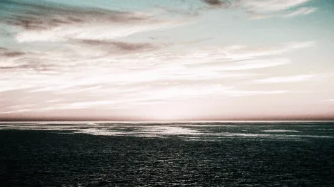 Breite Wolkenschichten über einer dunklen Meeresfläche, am Horizont leuchtet ein heller Sonnenuntergang in Rosa- und Blautönen.