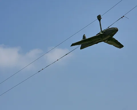 Dunkelgrünes Spielzeugflugzeug hängt schräg an Drähten vor blauem Himmel mit einer kleinen Wolke links.