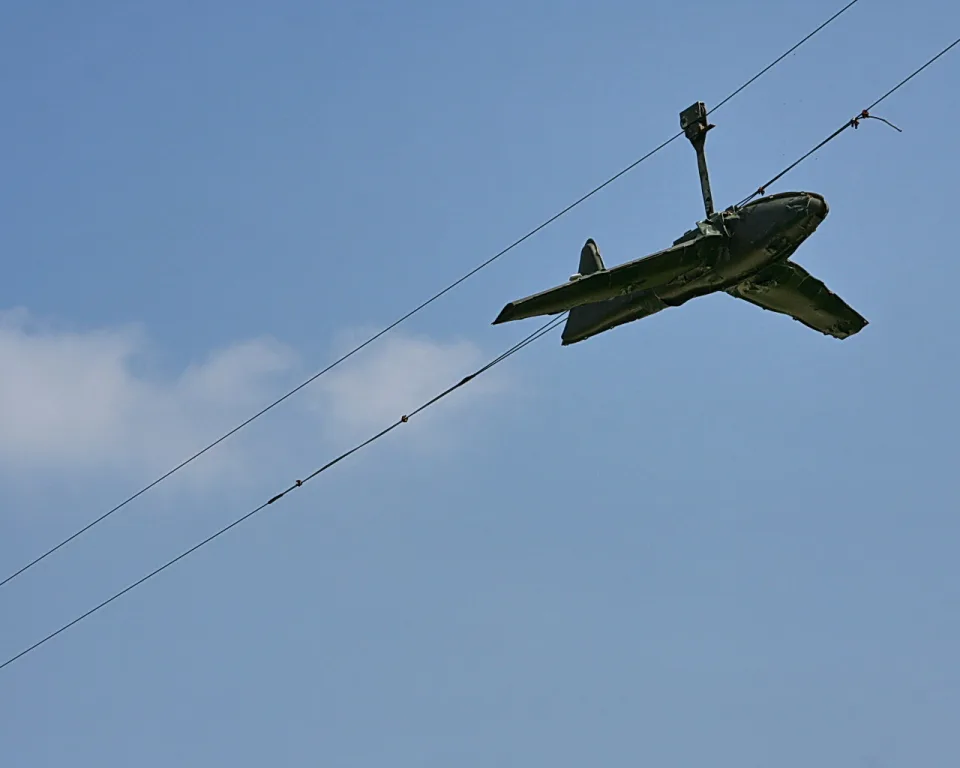 Dunkelgrünes Spielzeugflugzeug hängt schräg an Drähten vor blauem Himmel mit einer kleinen Wolke links.