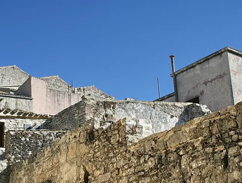 Alte, verwitterte Steinmauern und Hausdächer in einem sizilianischen Dorf unter wolkenlos blauem Himmel, rechts ein Gebäude mit Schornstein.