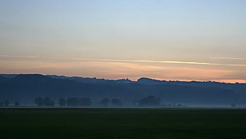 Weite Wiesenfläche im Vordergrund, dahinter eine dunkle Hügelkette im Dunst, darüber ein blau-lila Abendhimmel mit orangefarbenem Horizontstreifen und feinen Kondensstreifen.