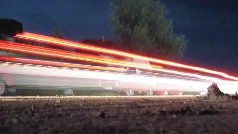 Rote und weiße Lichtspuren eines vorbeifahrenden Fahrzeugs auf einer nächtlichen Straße, im Hintergrund ein Baum und dunkler Himmel.