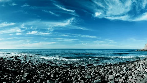 Breiter Strand aus dunklen, runden Kieselsteinen im Vordergrund, dahinter das blaue Meer mit Brandungswellen und ein Himmel mit feinen Schleierwolken; rechts am Rand ragt eine helle Felsklippe ins Bild.