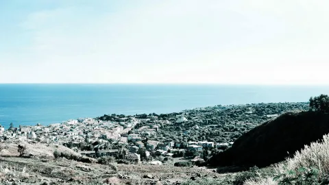 Kleine Küstenstadt mit weißen Häusern an einem Hang, dahinter das blaue Meer unter hellem Himmel, im Vordergrund trockenes Gras und ein dunkler Hügel.