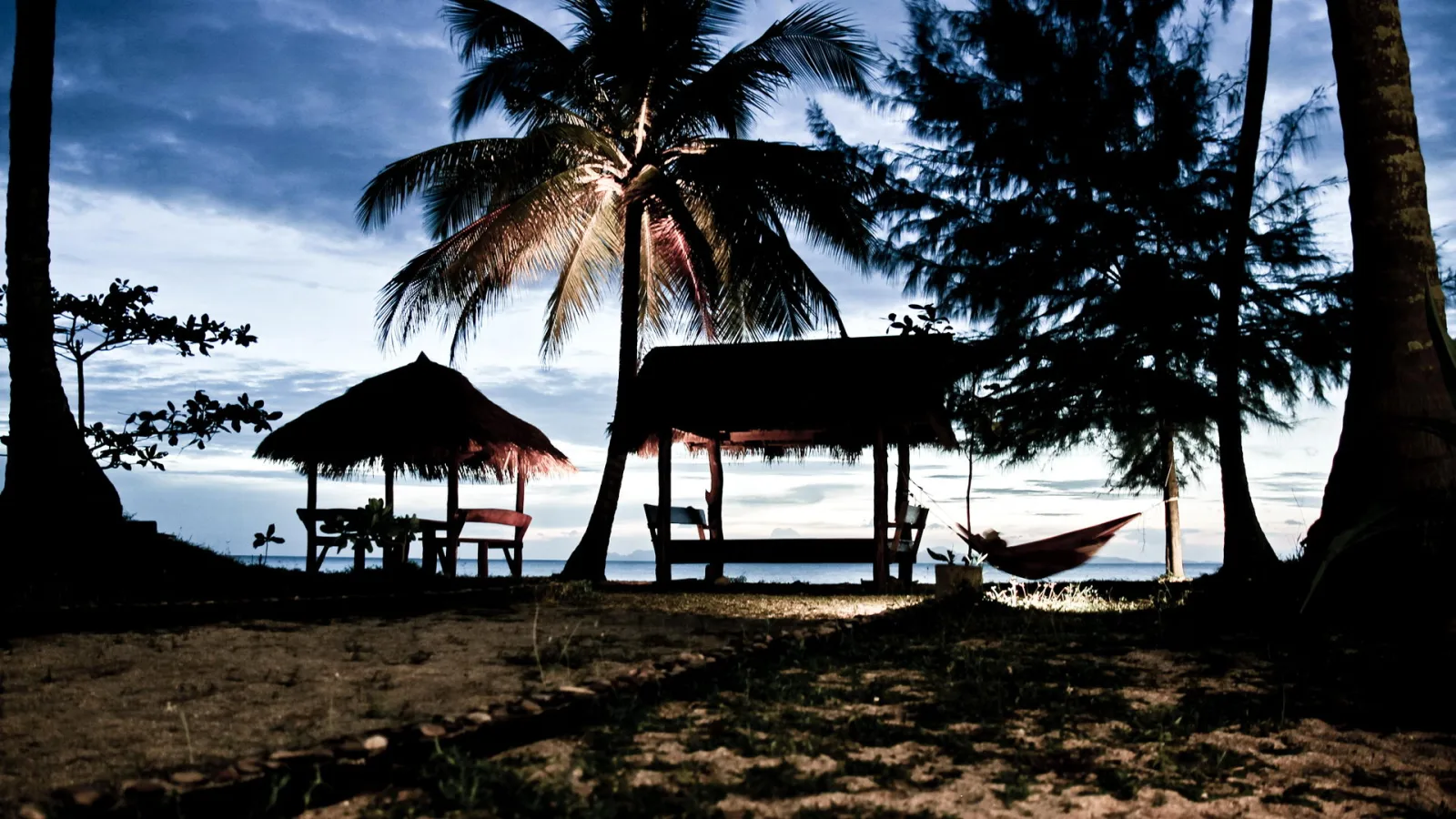 Strand am Abend mit zwei überdachten Holzunterständen, einer roten Hängematte zwischen Kiefern und Palmen vor dunkelblauem Himmel.