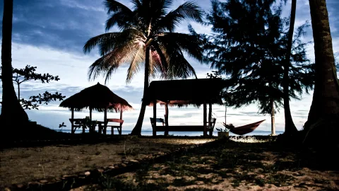 Strand am Abend mit zwei überdachten Holzunterständen, einer roten Hängematte zwischen Kiefern und Palmen vor dunkelblauem Himmel.