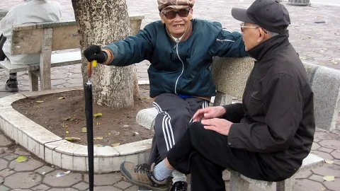 Zwei ältere Männer sitzen auf einer Steinbank neben einem Baum, der linke mit Sonnenbrille und Mütze zeigt mit einem Stock nach vorn, der rechte mit dunkler Kappe hört zu.