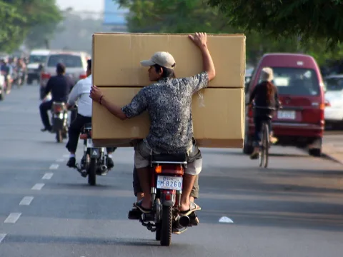 Ein Mann mit Basecap fährt auf einem roten Motorroller durch eine Straße und balanciert zwei große Kartons auf den Schultern, im Hintergrund weitere Motorroller und ein roter Minivan.