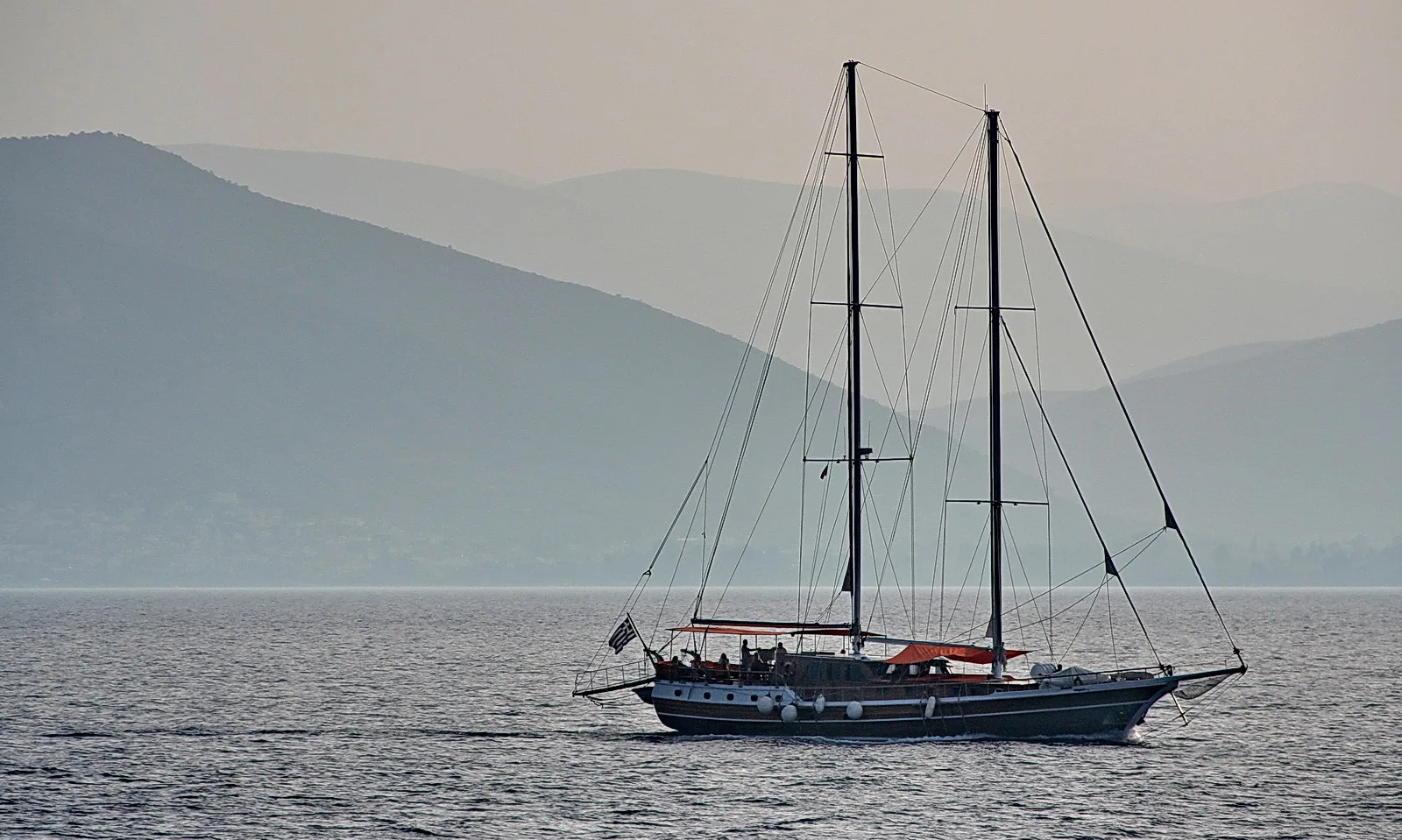 Eine zweimastige Segelyacht mit Dieselmotor und Takelage fährt auf ruhiger See, im Hintergrundliegen blaugraue Bergketten im Dunst.