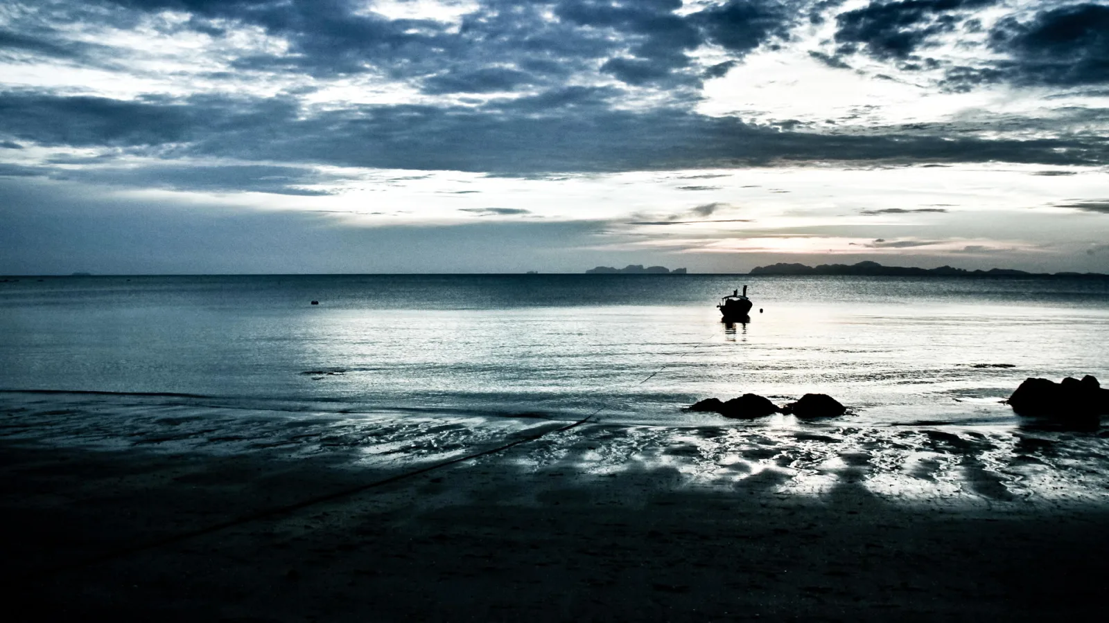 Ein kleines Boot als dunkle Silhouette auf ruhigem Meer, dahinter dunkle Wolken und ferne Inseln bei Dämmerung.