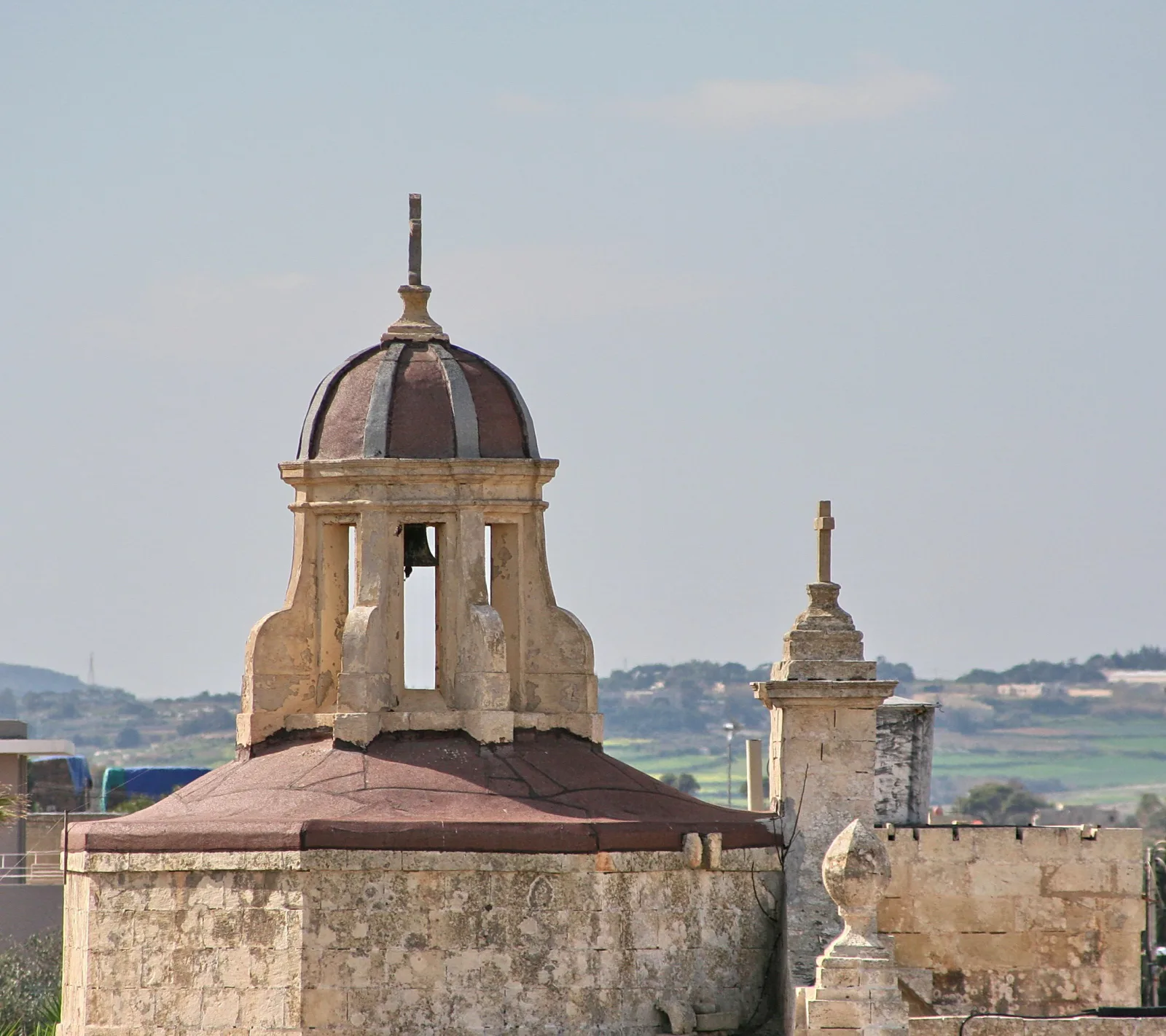 Kuppelförmiger Glockenturm aus hellem Kalkstein mit Glocke und Kreuz, daneben ein Pfeiler mit Kreuz, im Hintergrund Hügel unter diesigem Himmel.