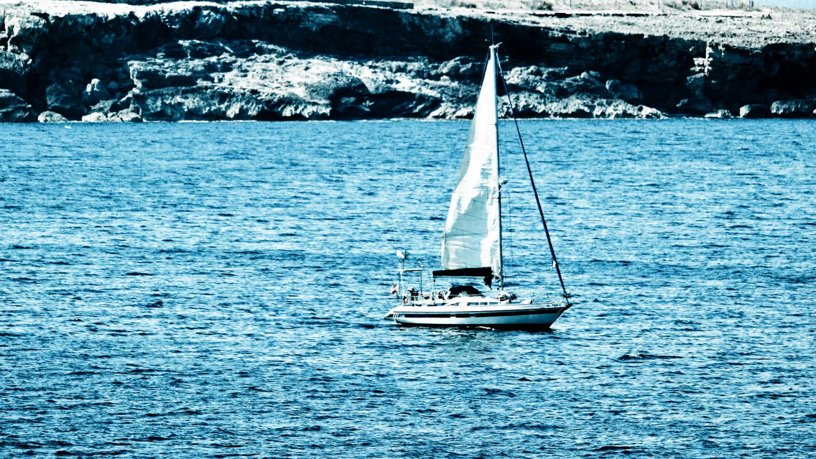Ein weißes Segelboot mit gesetztem Großsegel fährt auf einem blau getönten Meer vor einer felsigen Küste.
