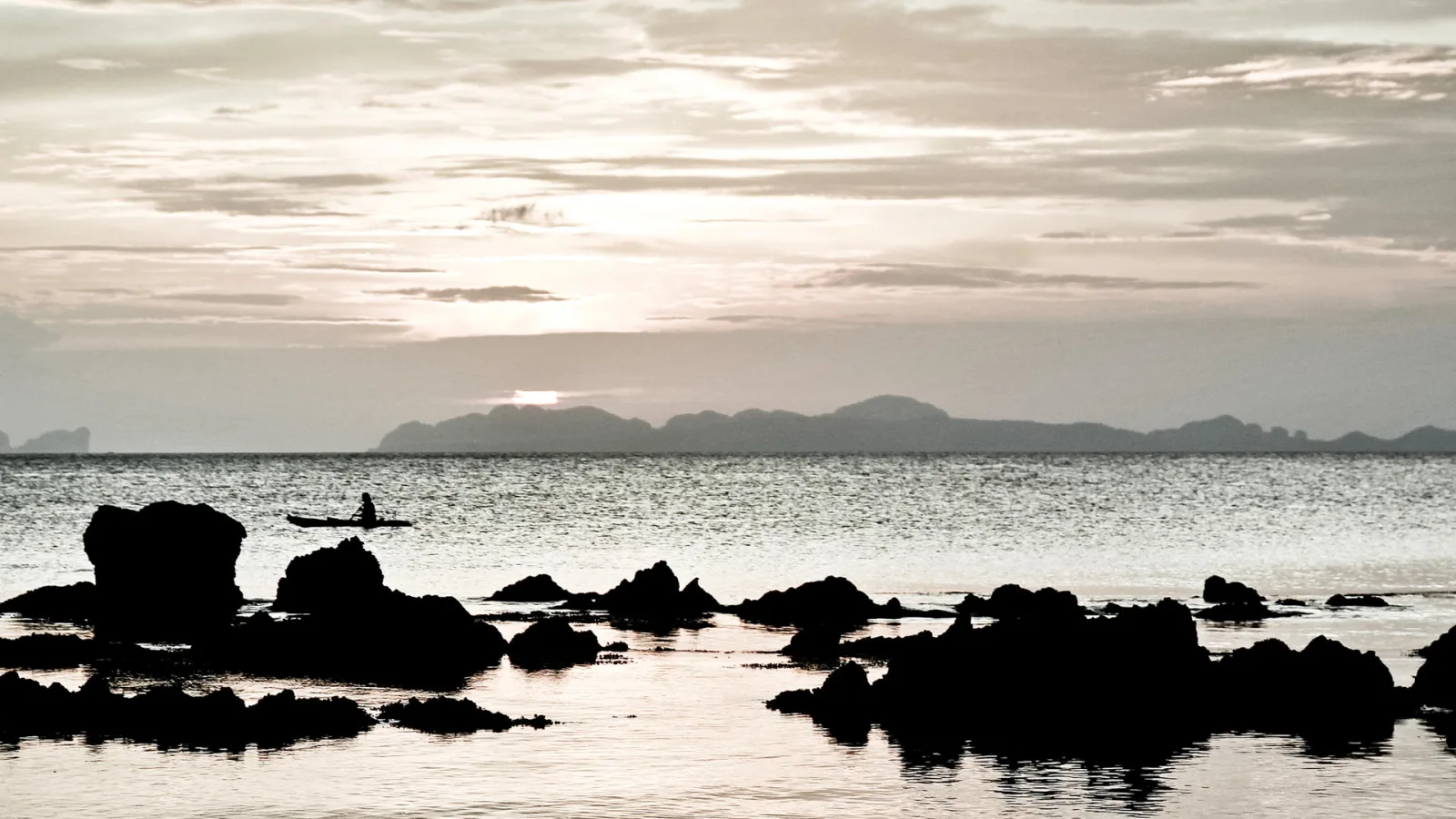 Silhouetten von Felsen im flachen Wasser, im Hintergrund ein Kajak mit einer Person und eine bergige Küstenlinie unter bewölktem Himmel bei Sonnenuntergang.