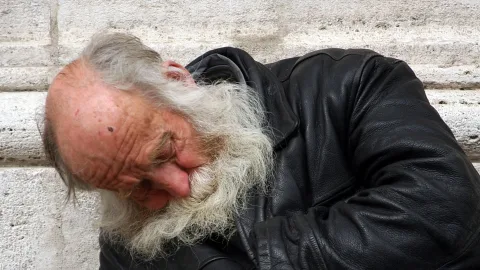 Älterer, kahlköpfiger Mann mit langem weißen Bart und schwarzer Lederjacke lehnt mit gesenktem Kopf an einer hellen Steinmauer.