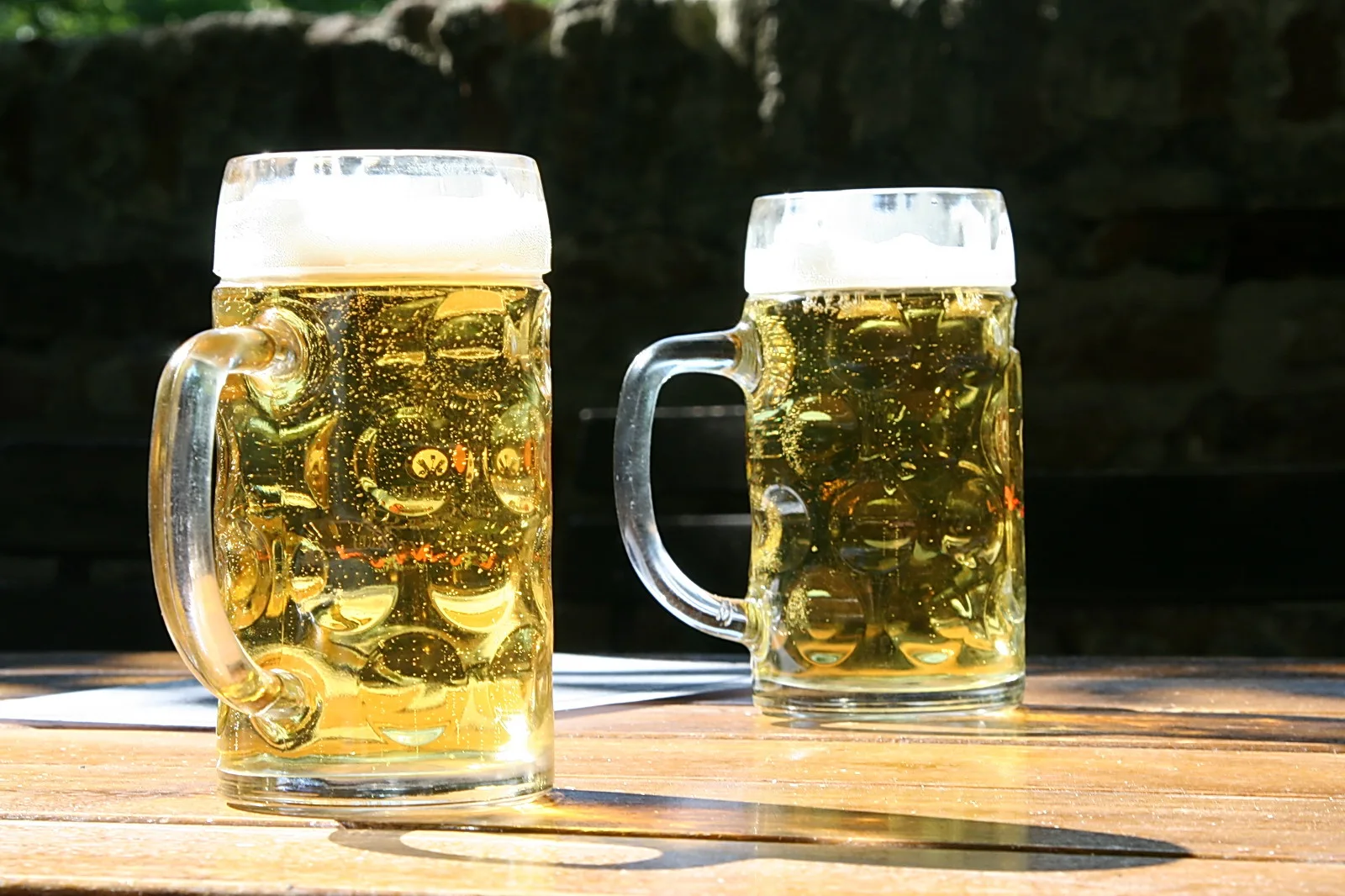 Zwei Bierkrüge mit Schaumkrone stehen im Sonnenlicht auf einem Holztisch, dahinter eine dunkle Steinmauer.