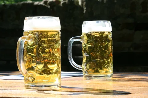 Zwei Bierkrüge mit Schaumkrone stehen im Sonnenlicht auf einem Holztisch, dahinter eine dunkle Steinmauer.