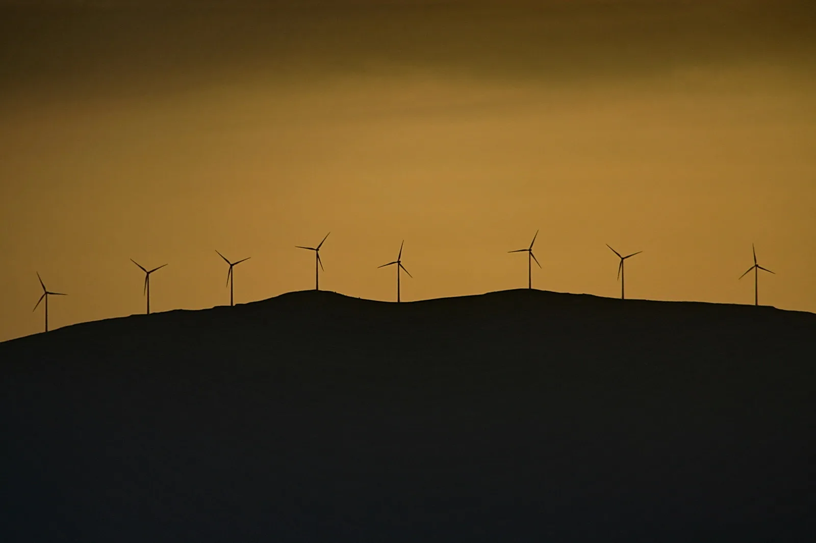 Eine Reihe von acht Windkraftanlagen zeichnet sich als Silhouetten auf einem dunklen Hügelkamm vor einem orangefarbenen Abendhimmel ab.
