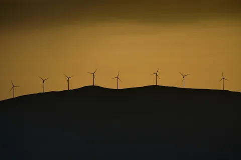 Eine Reihe von acht Windkraftanlagen zeichnet sich als Silhouetten auf einem dunklen Hügelkamm vor einem orangefarbenen Abendhimmel ab.