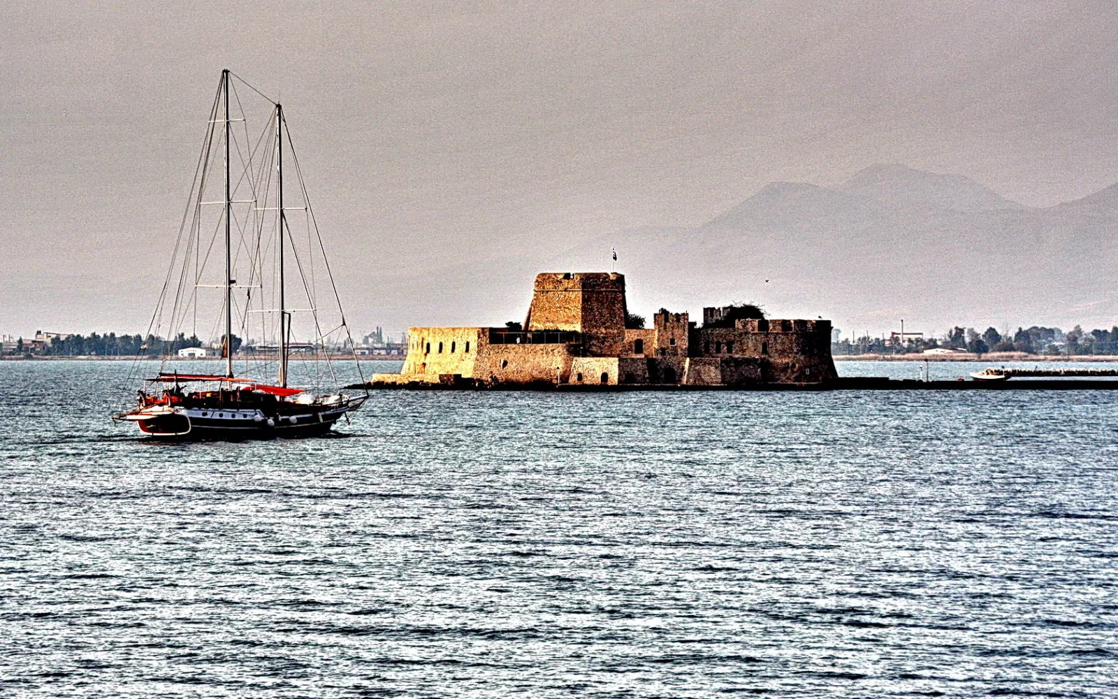 Eine alte Festung aus Stein steht auf einer Insel im Wasser, links liegt ein Segelschiff mit rotem Gaffelbaum vor grauem Himmel und Bergen.