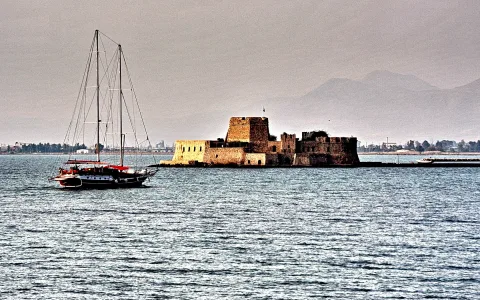 Eine alte Festung aus Stein steht auf einer Insel im Wasser, links liegt ein Segelschiff mit rotem Gaffelbaum vor grauem Himmel und Bergen.