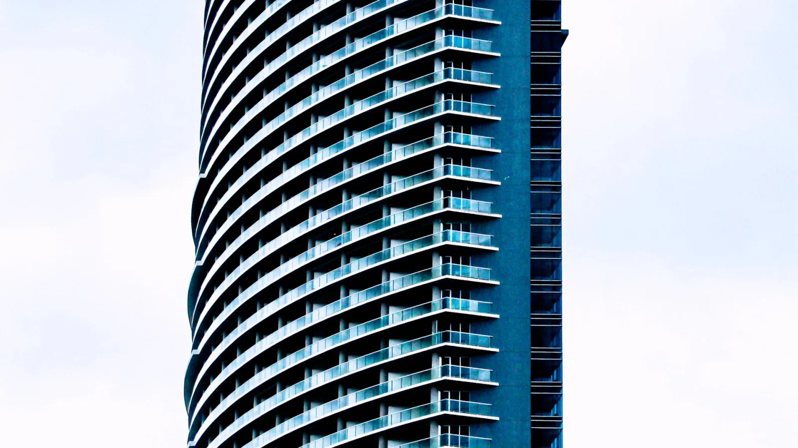 Schlanker Wolkenkratzer mit geschwungener Fassade und vielen übereinander gestaffelten Glasbalkonen vor hellgrauem Himmel