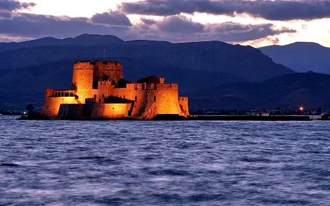 Beleuchtete Festung Bourtzi auf einer Insel bei Nafplio in der Abenddämmerung, davor dunkles Wasser, dahinter Bergketten unter einem bewölkten Himmel.