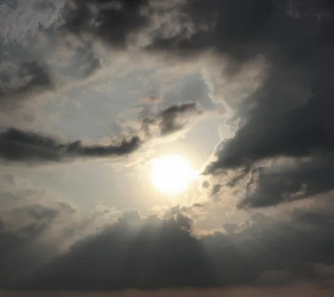 Helle Sonne bricht durch dunkle, aufgewühlte Wolken, von unten fallen Lichtstrahlen auf eine graue Wolkenbank.