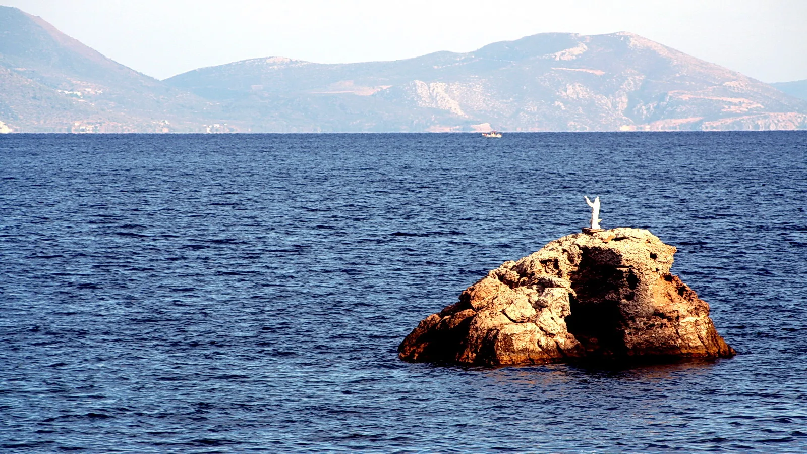 Weiße Jesusstatue mit erhobenen Armen auf einem Felsen im Meer, dahinter eine Bergkette im Dunst